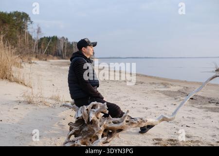 Seitenansicht eines Mannes, der auf Treibholz am Sandufer sitzt. Männlicher Reisender, der in der Nähe des Wassers ruht und den Horizont betrachtet. Das Konzept von Einsamkeit, Frieden und Natur. Stockfoto