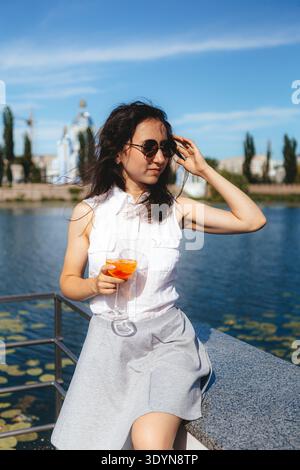 Eine Frau steht am Wasser und hält ein Glas mit einem Getränk. Sie trägt ein ärmelloses Hemd und einen Rock. Ein Park mit Bäumen ist im Hintergrund zu sehen Stockfoto