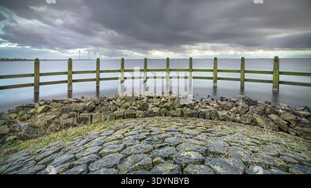 Der Holzzaun steht in den ruhigen Gewässern nahe Urk, mit entfernten Windturbinen vor einem dramatischen, bewölkten Himmel. Die Szene erfasst einen Stockfoto