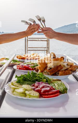 Eine Gruppe von Freunden genießt eine gemeinsame Mahlzeit auf einer Yacht und klickt in einer verspielten Geste über Tellern mit frischem Gemüse und Gebäck die Gabeln. Stockfoto
