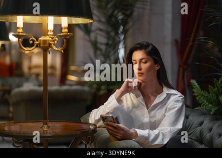 Junge Geschäftsfrau mit Smartphone, die auf gepolstertem Stuhl in der Nähe einer beleuchteten Lampe in der luxuriösen Hotellobby sitzt. Frau in weißem Hemd und wirkt entspannt Stockfoto