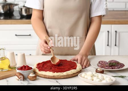 Frau mit Zutaten, die leckere Tomatensauce auf rohen Pizzateig in der Küche verteilt, Nahaufnahme Stockfoto