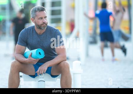Sport-Fitness-Mann, der draußen auf einer Bank ruht. Reifes Fitnesssportmodell, das nach dem Training auf einer Bank sitzt. Senior Sports man macht danach eine Pause Stockfoto