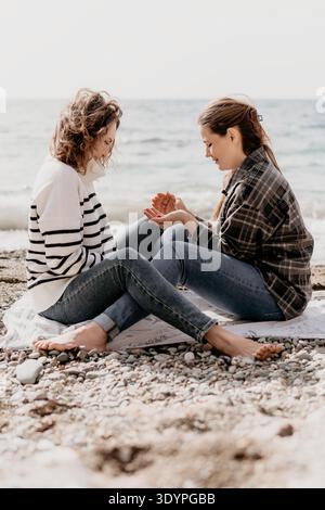 Strandfreundschaftsfrauen sitzen an einem sonnigen Tag am Meer zusammen an einem Kieselstrand und teilen ein kleines Fundstück Stockfoto