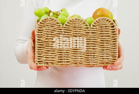 Frau mit einem gewebten Korb gefüllt mit frischen grünen Trauben und Birnen, Nahaufnahme der Hände, heller minimalistischer Hintergrund Stockfoto