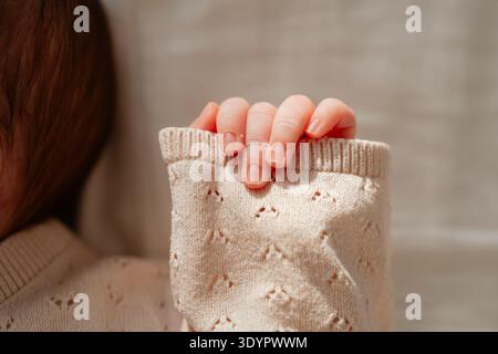 Nahaufnahme der Hand des Neugeborenen, die weiche Strickkleidung sanft hält. Warmes Sonnenlicht hebt zarte Finger und Textur hervor und symbolisiert Zärtlichkeit und Stockfoto