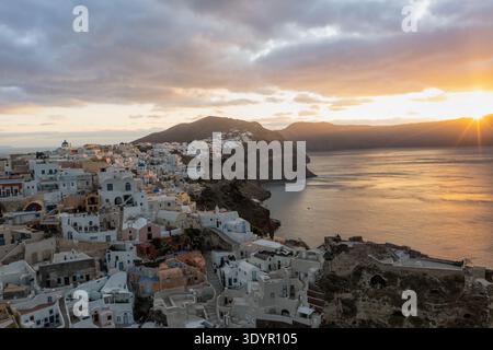Blick aus der Vogelperspektive auf weiße Gebäude kaskadieren Klippen in Richtung des türkisfarbenen Meeres, während die Sonne hinter den Hügeln, Fira, Thira, Griechenland, untergeht. Stockfoto