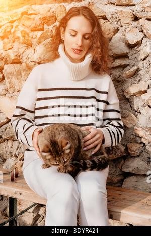 Frau Katze Haustier junge Frau in gestreiftem Rollkragenpullover sitzt vor der Felsenwand und streichelt eine gemütliche Tabby Katze auf ihrem Schoß Stockfoto