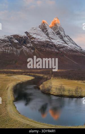 Aus der Vogelperspektive auf einen ruhigen Fluss, der sich durch goldene Felder unter den majestätischen, schneebedeckten Gipfeln der Berge schlängelt, deren Spitzen von der Wärme erstrahlen Stockfoto