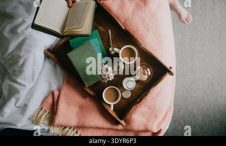 Auf einem Holztablett können Kaffee, Kerzen und Bücher auf einer gemütlichen rosa Decke aufbewahrt werden. Das offene Buch lädt zum Lesen oder zur Ruhe am Morgen ein. Das Tablett liegt neben dem weichen weißen Bett Stockfoto