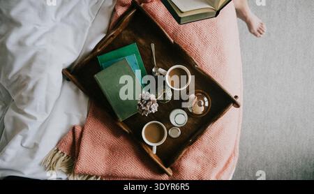 Auf einem Holztablett können Kaffeetassen, Bücher und Blumen auf einem gemütlichen Bett aufbewahrt werden. Weiche Decken und neutrale Töne schaffen eine ruhige, entspannende Atmosphäre. Die Szene deutet darauf hin Stockfoto