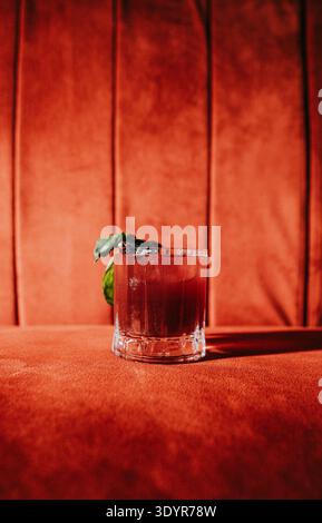 Kräftiger, dunkelroter Cocktail in einem Steinglas, garniert mit Minze und Limette. Es liegt auf einer strukturierten roten Oberfläche vor einem warmen, hölzernen Hintergrund. Die Lichtquelle Stockfoto
