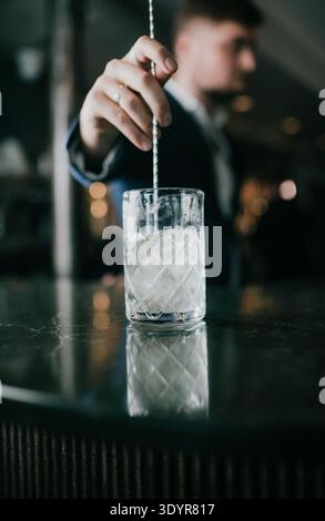 Barkeeper rührt Cocktail mit langem Rührer in klarem Glas. Eiswürfel funkeln unter der warmen Barbeleuchtung. Der Barkeeper trägt einen dunklen Anzug und einen fokussierten Ausdruck. D Stockfoto