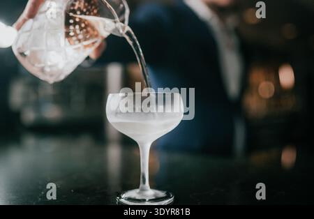 Cocktailglas wird mit klarer Flüssigkeit aus dem Jigger gefüllt. Der Barkeeper gießt Getränke mit Präzision. Das Glas liegt auf einer dunklen Staboberfläche. Hintergrund unscharf wi Stockfoto