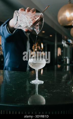 Gießt klare Flüssigkeit von Hand in Cocktailglas. Der Barkeeper in blauem Hemd serviert Getränke. Das Glas sitzt auf einer dunklen, glänzenden Staboberfläche. Warme Beleuchtung hebt po Stockfoto