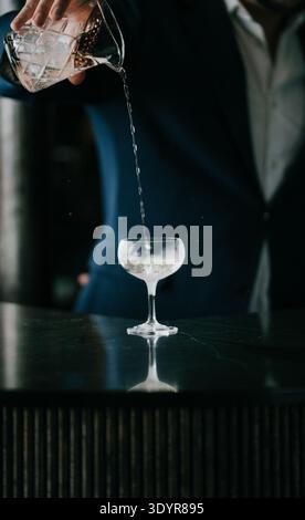 Gießt klare Flüssigkeit von Hand in Cocktailglas. Der Barkeeper in Anzug bereitet Getränke mit Sorgfalt zu. Dunkler Hintergrund hebt Glas hervor und gießt. Perfekt für Bar, n Stockfoto