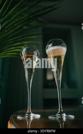 Zwei Sektflöten, gefüllt mit sprudelnder goldener Flüssigkeit, sitzen auf einem dunklen Holztisch. Sanfte, warme Beleuchtung unterstreicht die eleganten Kurven und den Schimmer der Brille Stockfoto