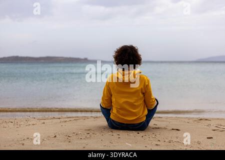 Rückansicht einer Frau in gelbem Hoodie, die in Lotusposition am Strand sitzt Stockfoto