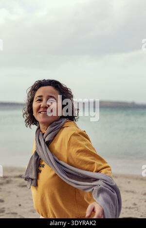 Frau mittleren Alters im Kapuzenpullover und grauen Schal lächelt am Sandstrand Stockfoto