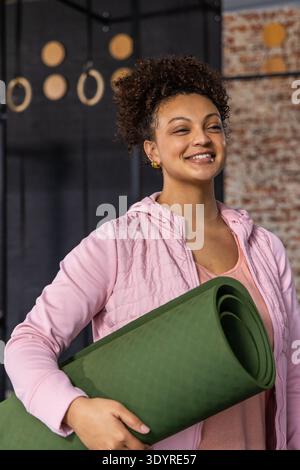 Afroamerikanerin, 20iger, die eine grüne Yogamatte im Fitness Studio trägt und eine rosa Jacke trägt Stockfoto