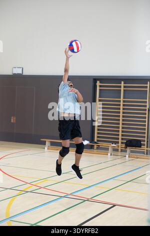 Afroamerikanischer Teenager, der graues T-Shirt trägt, springt und greift in der Turnhalle, um Volleyball zu schlagen. Athlet, Jugend, Midair, Ball, Bars, Bank, Sporthalle || Modell veröffentlicht Stockfoto