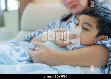 Mutter misst die Temperatur ihres kranken Kindes. Krankes Kind mit hohem Fieber, Mutter mit Thermometer und Tuch zur Nasenreinigung Stockfoto