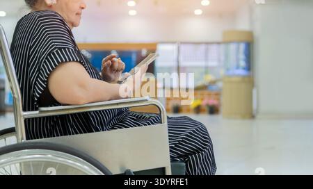 Seitenansicht einer asiatischen älteren Frau Patient benutzen Handy während sitzend und glücklich auf Rollstuhl im Krankenhaus mit Kopierraum. Selektiver Fokus. Stockfoto