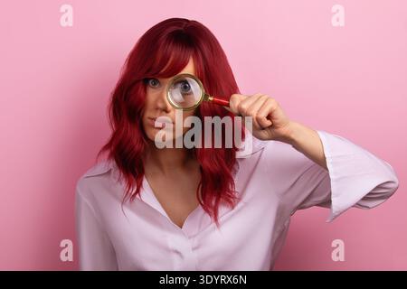 Junge Frau mit rotem Haar, die durch eine Lupe untersucht und vor einem pastellrosa Hintergrund posiert. Stockfoto