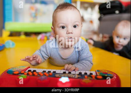 Ein Baby liegt auf einer gelben Matte und interagiert mit einem roten Tastaturspielzeug. Ein weiteres Baby ist im Hintergrund zu sehen. Die Umgebung ist ein helles Spielzimmer gefüllt Stockfoto