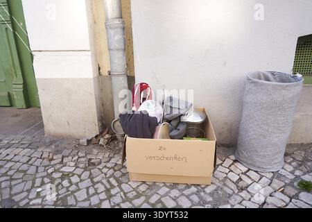 Zum Verschenken befindet sich eine Schachtel mit weggeworfenen Gegenständen auf der Straße vor der Tür, Karton mit der Aufschrift „to give off“ und Kleidung neben einer Seite Stockfoto