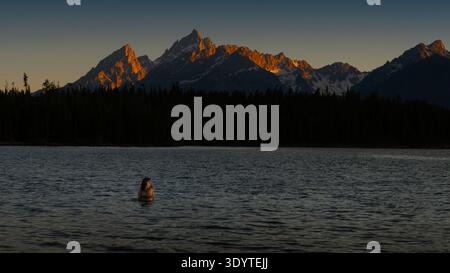 Ein Paar, das sich in einem See unter schneebedeckten Bergen umgibt und den Sonnenuntergang beobachtet. Stockfoto