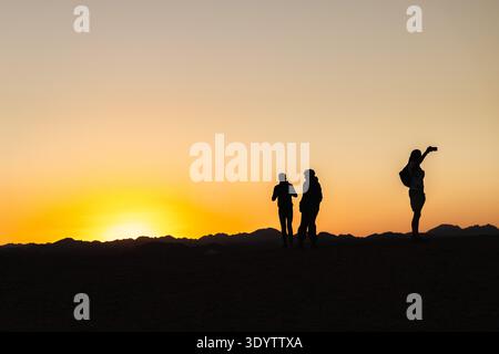 Silhouetten von Touristen, die den Sonnenuntergang in der ägyptischen Wüste mit Bergen am Horizont beobachten Stockfoto