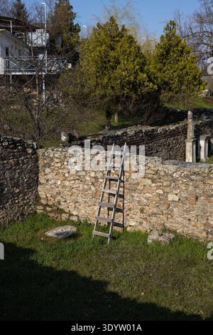Holzleiter, die an einer verwitterten alten Steinmauer in einer sonnendurchfluteten archäologischen Stätte mit rustikalen Texturen, üppigem Grün und historischen Ruinen anlehnt Stockfoto