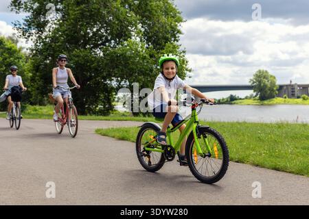 Familienradeln auf dem Radweg am Fluss nahe der Elbe in Magdeburg unter dramatischen Wolken. Mutter Kind, das Fahrrad-Helme durch grüne Landschaft reitet Stockfoto