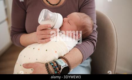Die Frau Füttert Das Neugeborene Mit Einer Flasche, Während Sie Das Baby Während Des Ruhigen Nachmittags Zu Hause In Ein Tuch Gewickelt Hält. Stockfoto