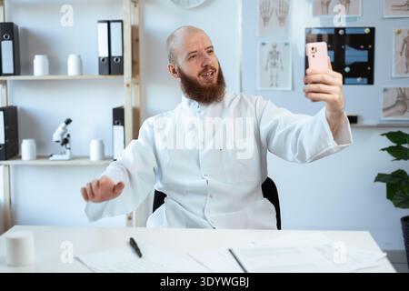 Lächelnder bärtiger männlicher Arzt mit Rezepten, Medikamentendokumenten Selfie machen funktioniert im Krankenhausbüro. Kopierbereich Stockfoto