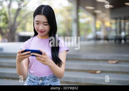 Junge Frau lächelt gerne draußen digitale Unterhaltung. Sie nutzt ein Handheld-Gerät, um an einem hellen Tag Inhalte zu durchsuchen. Stockfoto