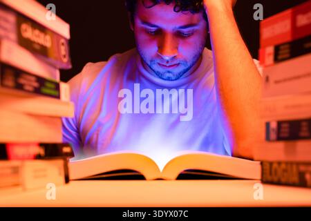 Junger Mann, der zwischen Büchern in seinem Zimmer mit Licht aus dem Buch studiert und auf seine Lektüre konzentriert sitzt Stockfoto