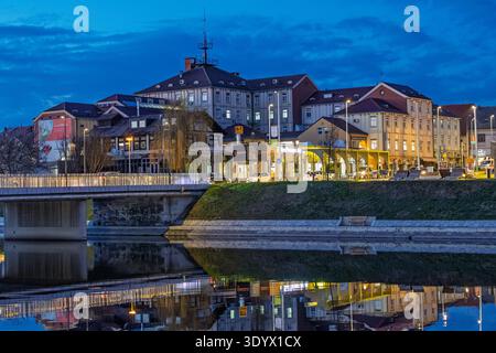 VINKOVCI, KROATIEN - 18. Februar 2026: Ruhiger Abendblick auf Gebäude am Fluss und Bäume, die sich in einem ruhigen Fluss spiegeln und von warmen Straßenlichtern beleuchtet werden Stockfoto