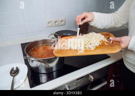Weibliche Hände geben frischen Kohl aus einem Holzbrett in einen Metalltopf mit traditionellem ukrainischem Borschtsch auf einem modernen Herd in der Küche Stockfoto