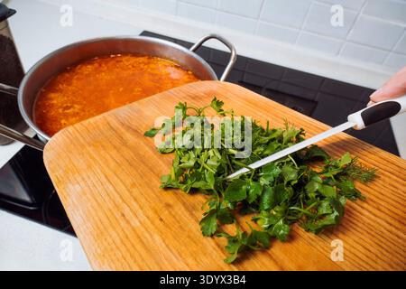 Kochen Sie frische grüne Petersilie und Dill von einem Holzbrett in einen Topf mit traditionellem ukrainischem Borschtsch, um den Geschmack der gesunden Suppe zu verstärken Stockfoto