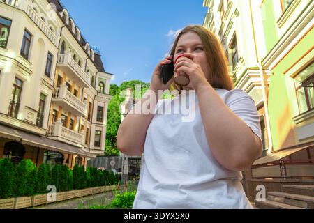 Junge Frau in einem weißen T-Shirt, die am Telefon spricht und Kaffee zum Mitnehmen trinkt, auf einer charmanten europäischen Stadtstraße mit wunderschönen Gebäuden Stockfoto