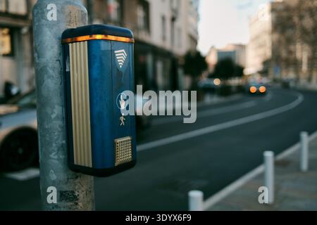 Fußgängerüberquerungsanforderungstaste, die an einem Metallpfosten neben einer städtischen Straße montiert ist, mit Autos und Gebäuden im Hintergrund verschwommen in Belgrad. Stockfoto