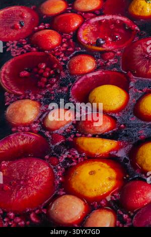 Nahaufnahme von Zitrusscheiben, Äpfeln und gemischten Beeren, die in dunklem Fruchtpunsch schweben, wodurch leuchtende rote und gelbe Kontraste und reichhaltige Fruchttexturen entstehen. Stockfoto