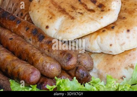 Nahaufnahme gegrillter Würstchen mit warmem Pita-Brot und frischem Salat, die als rustikales Grillgericht im Freien mit reichhaltigen Grilltexturen serviert werden. Stockfoto