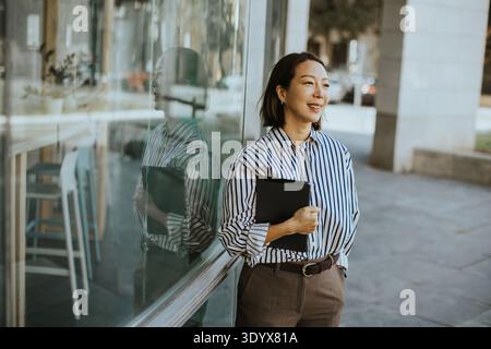 Eine Koreanerin steht nachmittags vor einem Café. Sie hält eine schwarze Mappe und lächelt, während sie hinausschaut. Die Sonne reflektiert auf dem Fenster Stockfoto