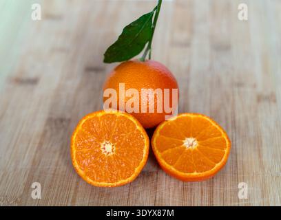 Frische reife Mandarine mit grünem Blatt und zwei geschnittenen Hälften auf hölzernem Hintergrund. Gesunde Zitrusfrucht reich an Vitamin C und natürlichem Antioxidans Stockfoto