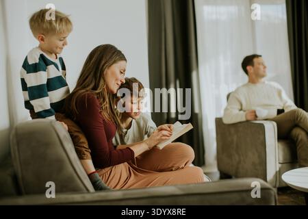 Zwei Kinder und eine Frau lasen ein Buch in einem gemütlichen Wohnzimmer. Ein Mann sitzt auf einem anderen Stuhl und genießt eine Tasse Kaffee. Die Sonne scheint durch die Fenster Stockfoto