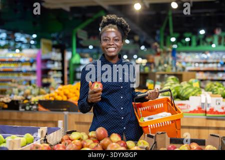 Glückliche Frau, die einen Reifen roten Apfel und einen orangen Warenkorb hält, während sie im Lebensmittelbereich steht und gesunde Früchte für einen nahrhaften Lebensstil auswählt Stockfoto