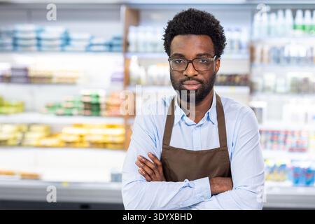 Professioneller Mitarbeiter im Einzelhandel mit Brille und Schürze, der mit überkreuzten Armen vor verschwommenen Regalen in Supermärkten steht, steht für Kundenservice und engagierte Einstellung am Arbeitsplatz Stockfoto